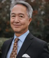 Smiling man in a suit and patterned tie, standing outdoors with trees in the background.