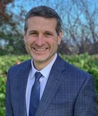 Smiling man in a blue suit and tie standing outdoors with greenery and trees behind him.