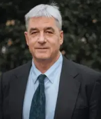 Man with short gray hair in a suit and tie standing outdoors in front of greenery.