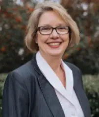 Smiling woman with short blonde hair, glasses, and a suit jacket, outdoors.
