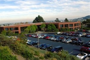 A large office building with a parking lot full of cars and trees in the background.