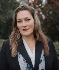 Woman with wavy hair in a suit jacket standing outdoors with trees in the background.