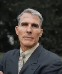 A serious man in a suit stands outdoors with trees in the blurred background.