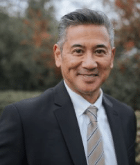 Smiling man in a suit and tie standing outdoors with blurred trees in the background.