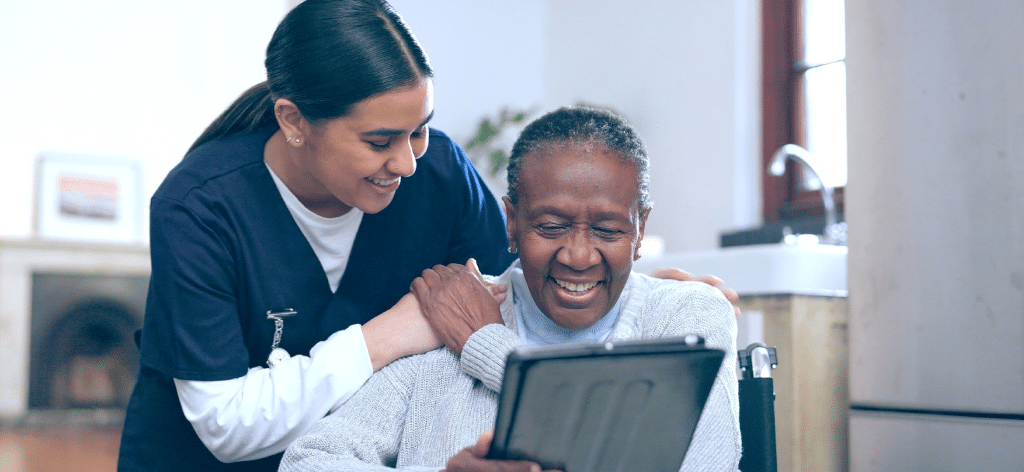 About InClin: Smiling nurse helps elderly woman use a tablet in a bright, cozy room.